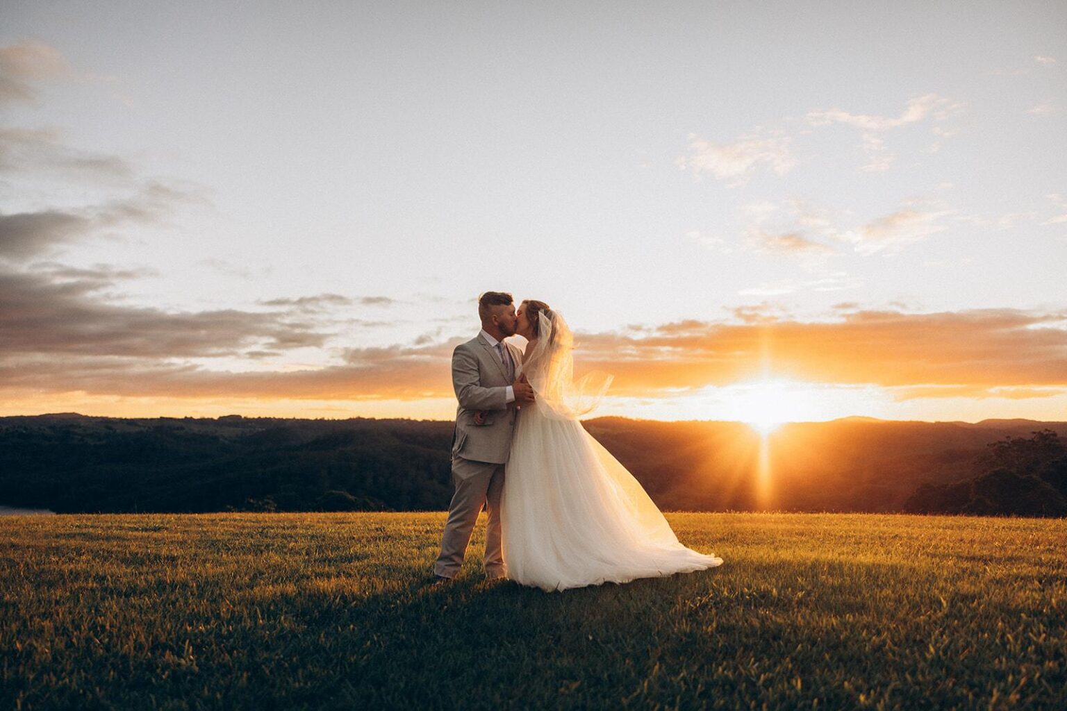 Bride and groom in a white wedding dress and light suit kissing in a sunlit field at sunset.