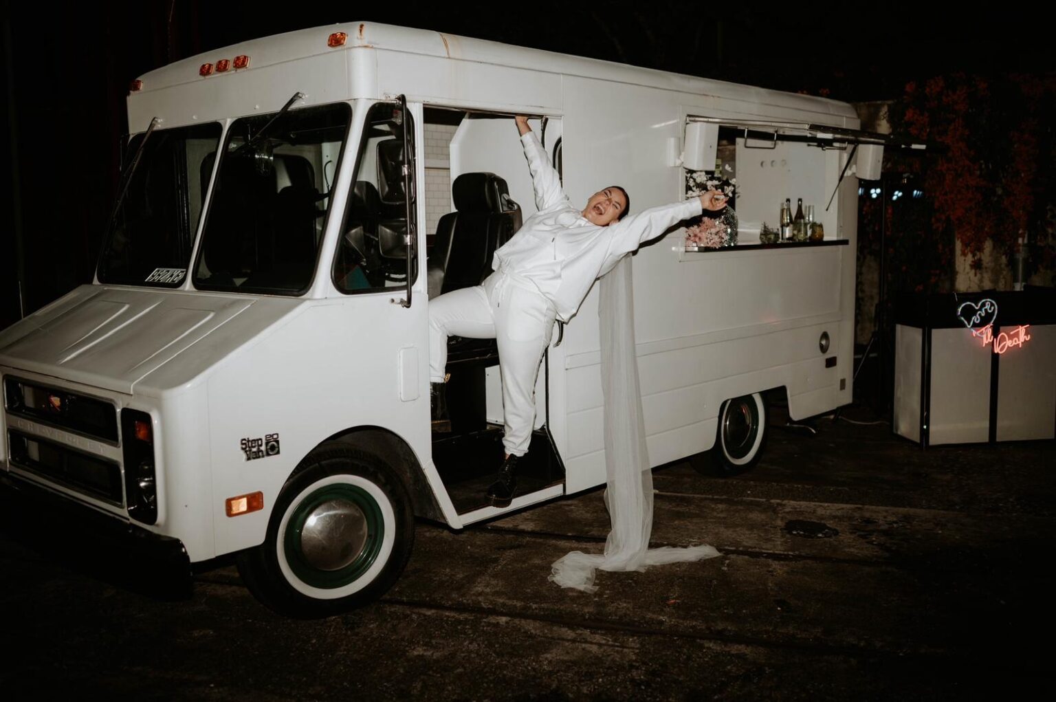 Groom in a white suit leans from an open vintage bus at night, with a long veil trailing behind him.