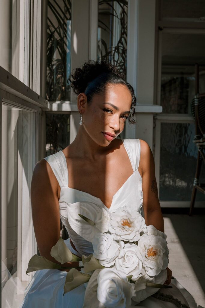 Woman in a white wedding dress stands in a sunlit doorway, holding a bouquet of white blooms closer to her waist.