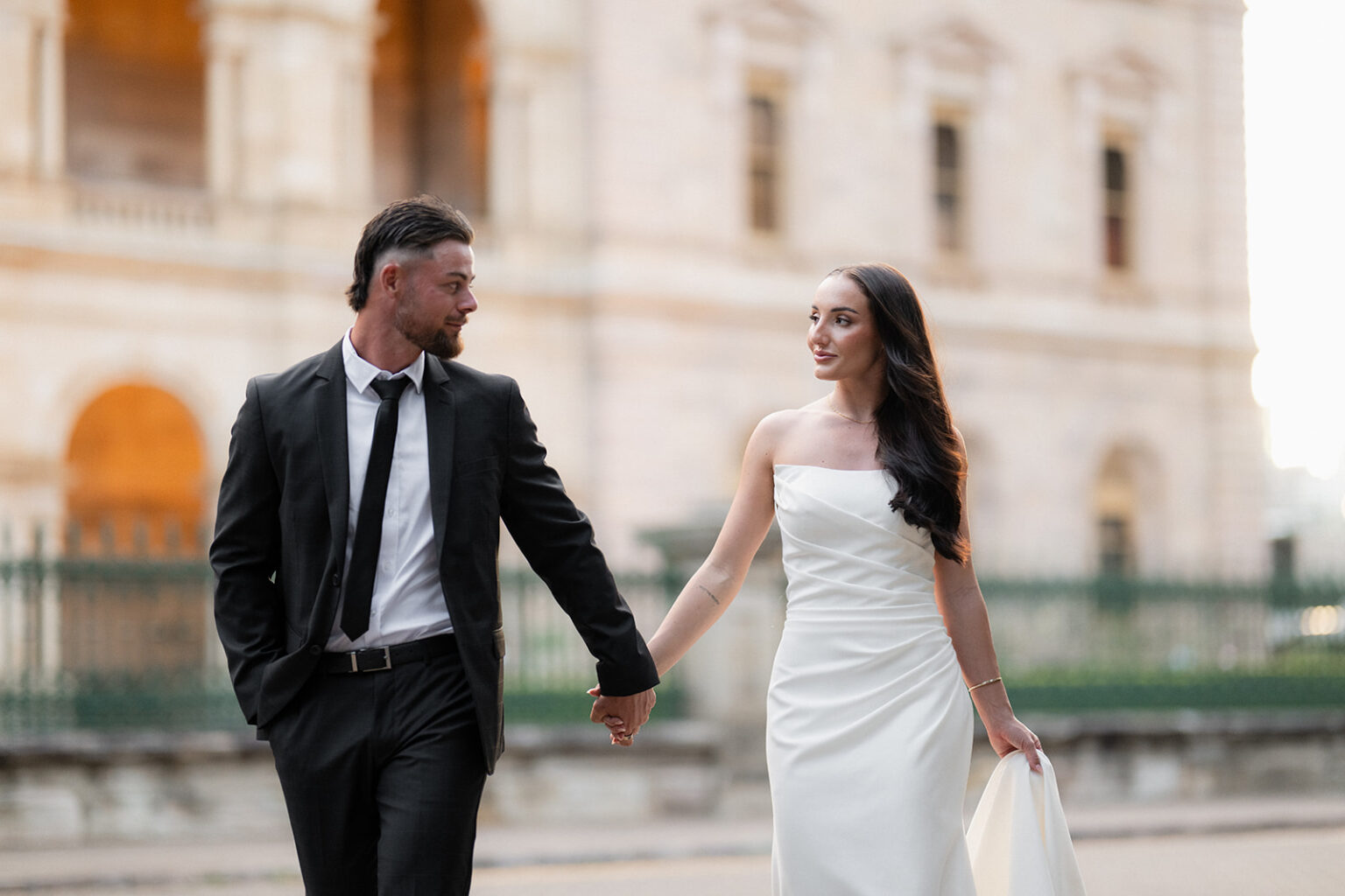 A bride in a white strapless wedding gown and a groom in a black suit walk hand in hand outdoors near a stone building at sunset.