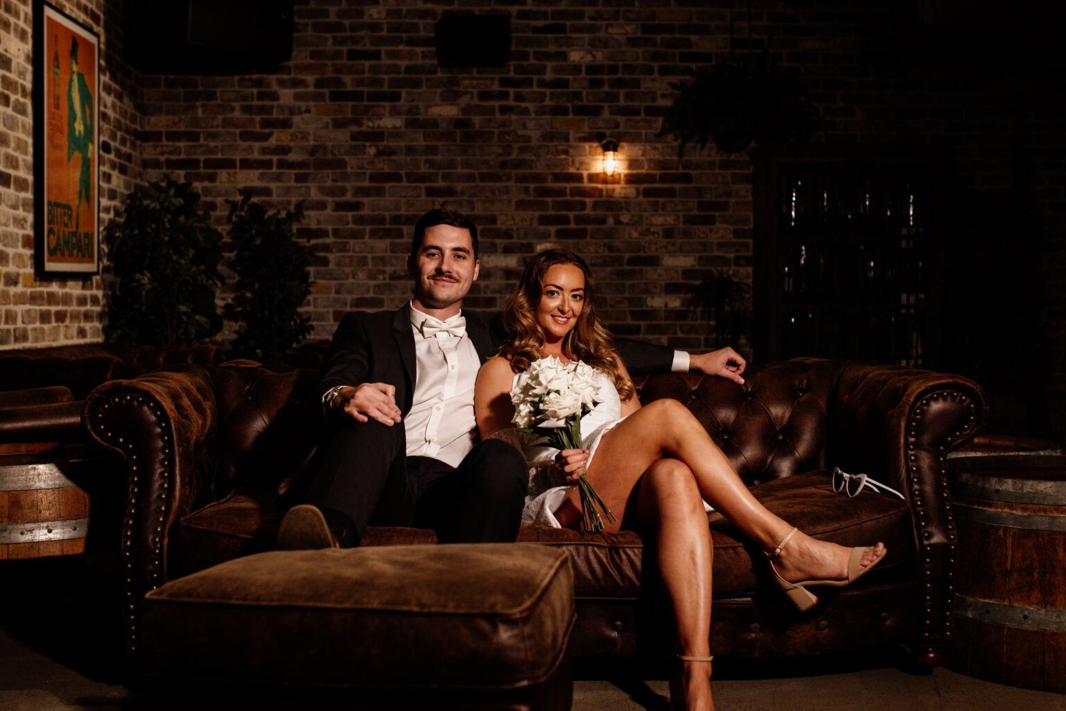Couple in formal wear sitting on a brown leather sofa in a dim brick-walled lounge; the woman holds a bouquet and smiles at the camera.